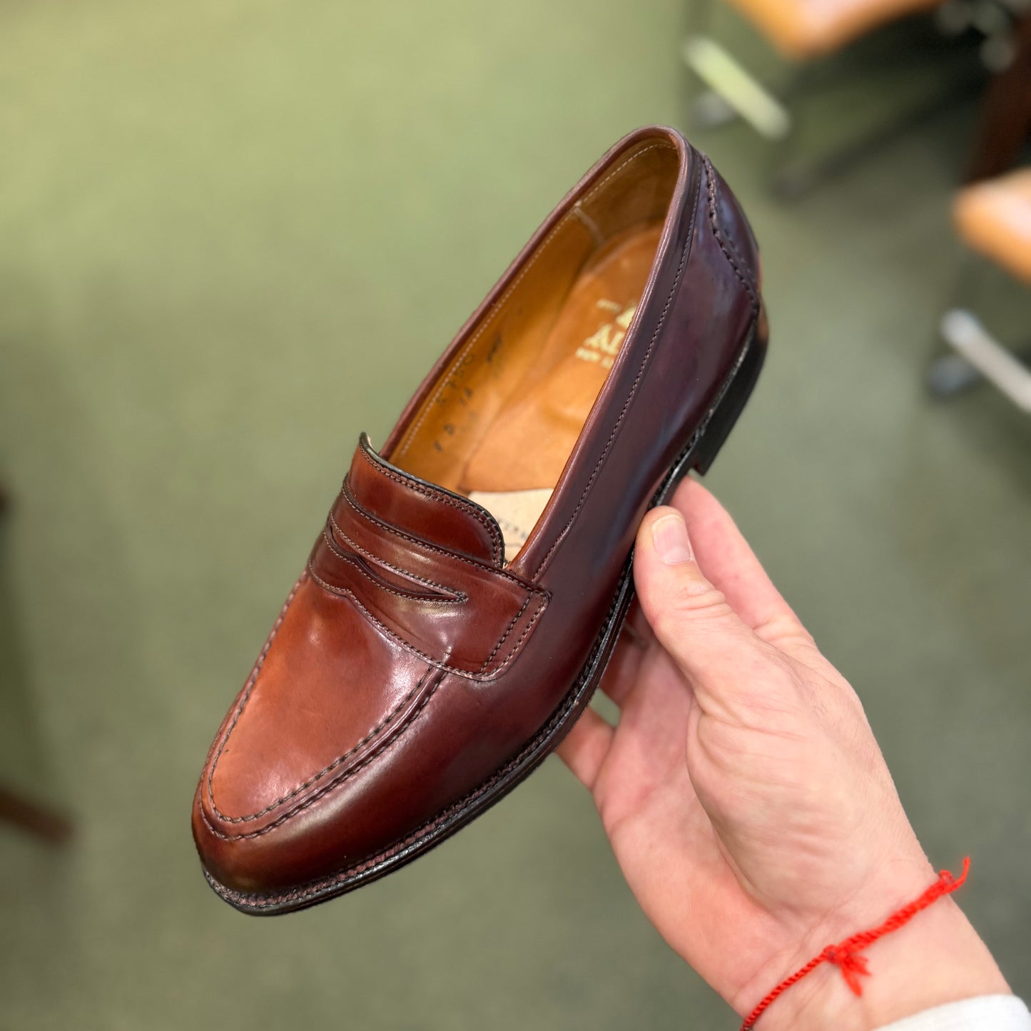 A hand holds the ALDEN SHOES 5750 - Higher Vamp "Midtown" Slip On in Color 8 above a green carpeted floor, with blurred chairs in the background.