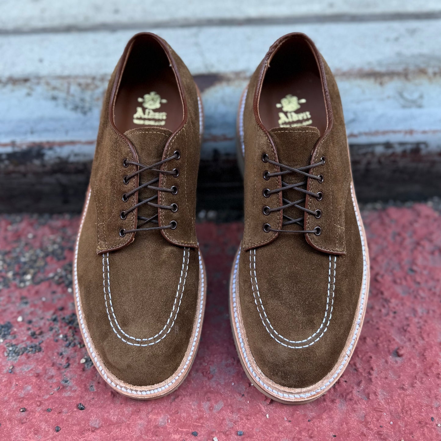 A pair of ALDEN SHOES M7601Y - Snuff Suede Indy Shoes with visible white stitching and brown laces rests on a surface shifting from red to gray, revealing the brand logo and highlighting their distinctive Trubalance Last design.