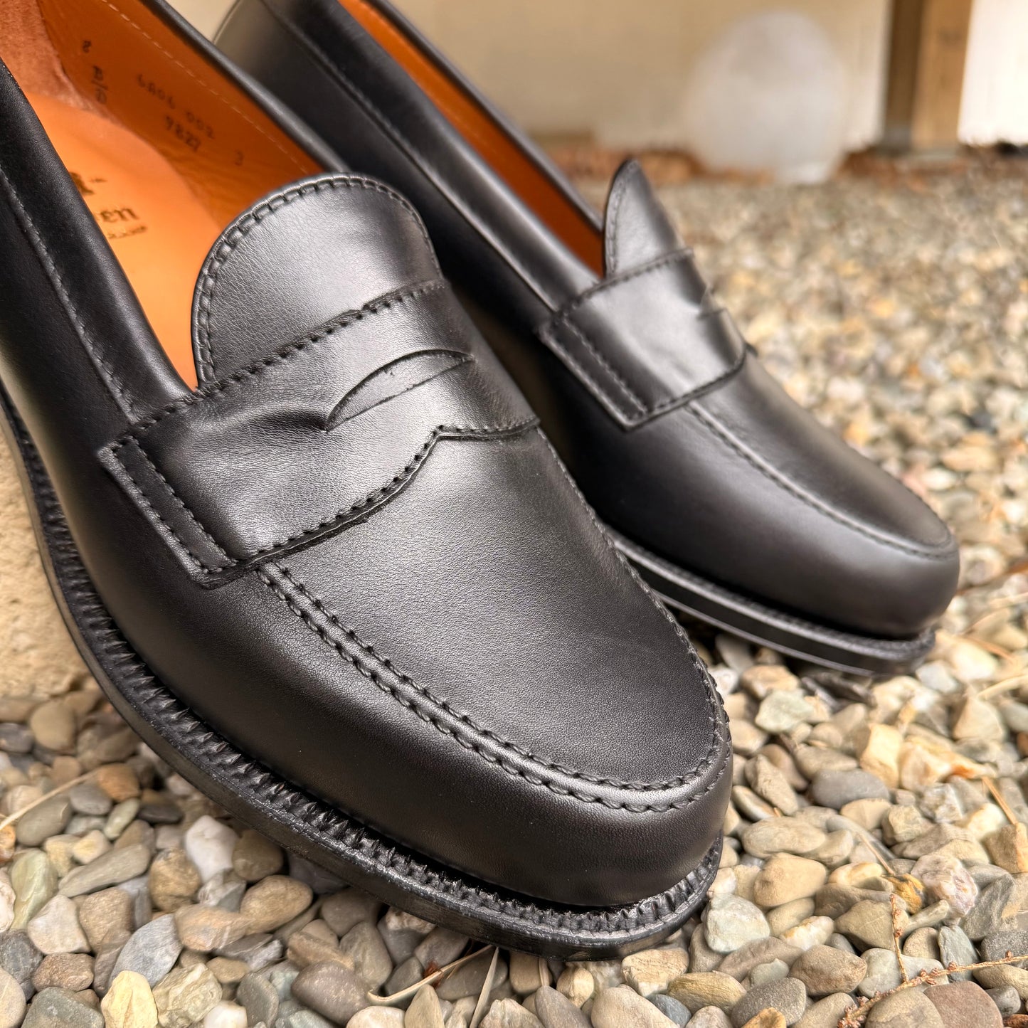 Close-up of ALDEN SHOES 9827 - Black Calf Leisure Penny Loafer, featuring visible stitching and a single leather sole, displayed on small pebbles with a softly blurred background.