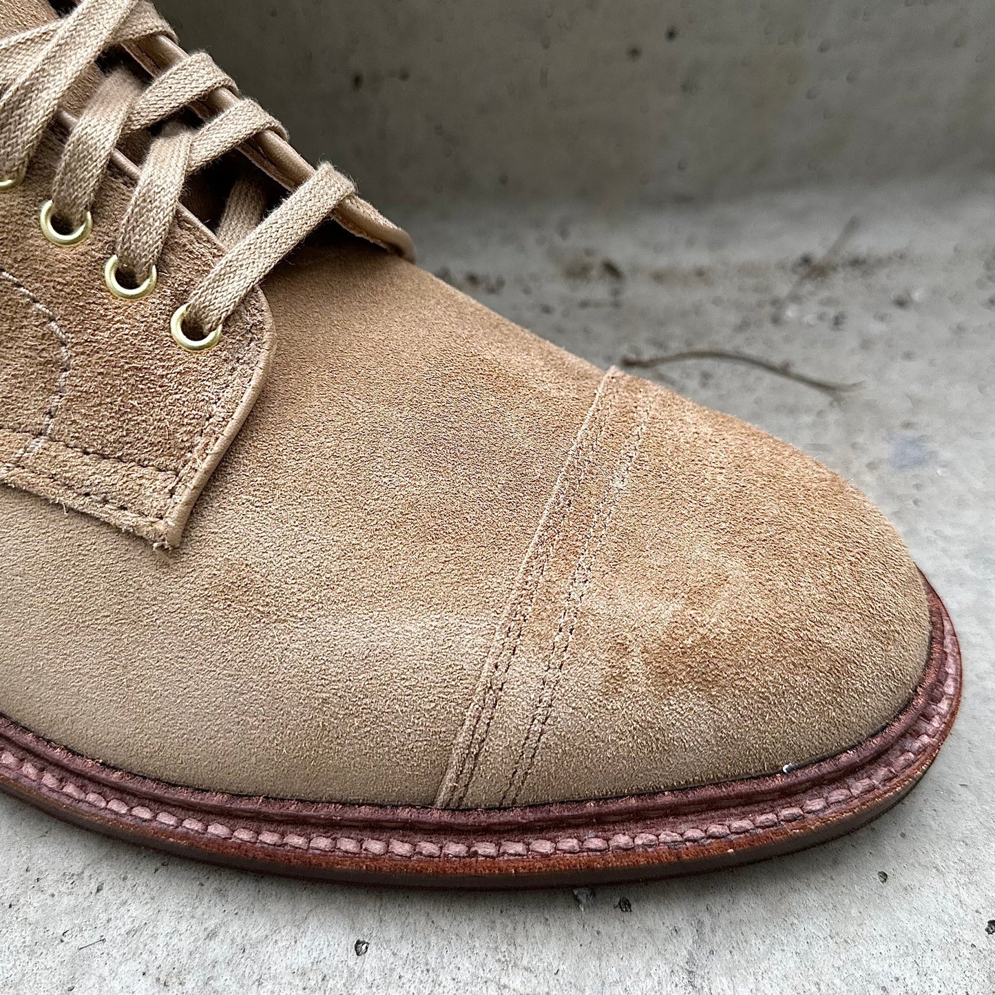 Close-up of the Alden Madison D2685H - 2/2 Cap Toe Boot in tan suede, featuring gold eyelets and beige laces, crafted on the Barrie Last. Detailed stitching accents the toe cap and sole as it rests elegantly on a textured concrete surface.