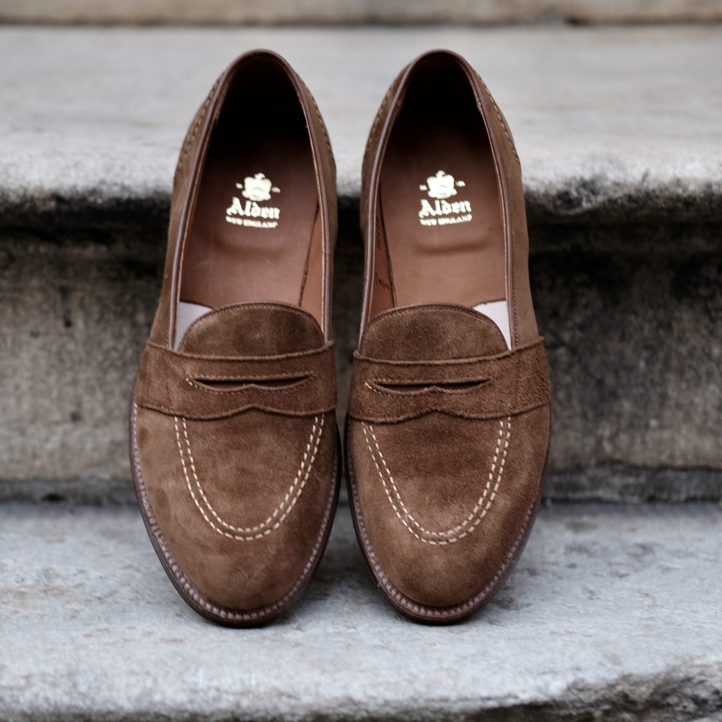 Alden Madison’s D0109 - Full Strap Loafer in snuff suede, featuring white stitching and leather outsoles, displayed side by side on stone steps. Gold Alden logo details the insoles.