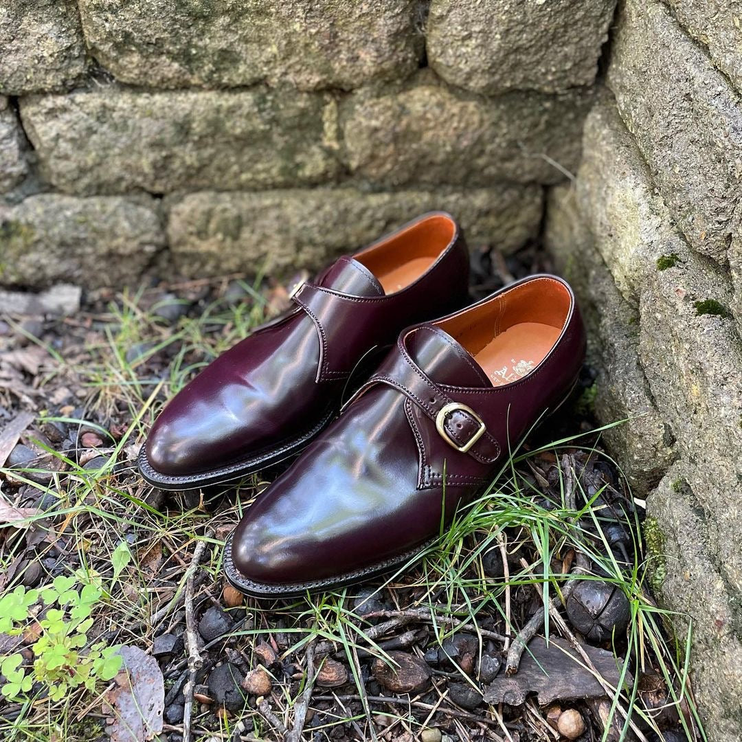 A pair of Alden Madisons 954 - Monk Strap Color 8 Shell Cordovan shoes, elegantly resting by a stone wall with fallen leaves and green plants surrounding them.