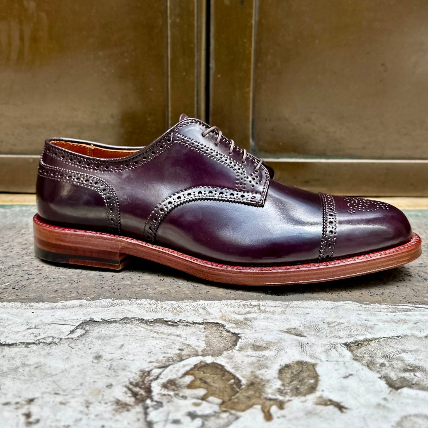 The Alden Madison 21522 - Medallion Cap Toe Blucher Color 8, crafted from Shell Cordovan leather, showcases elegant and durable dark brown polished leather with intricate brogue detailing on the toe cap and sides, against a metallic bronze background on a textured concrete surface.