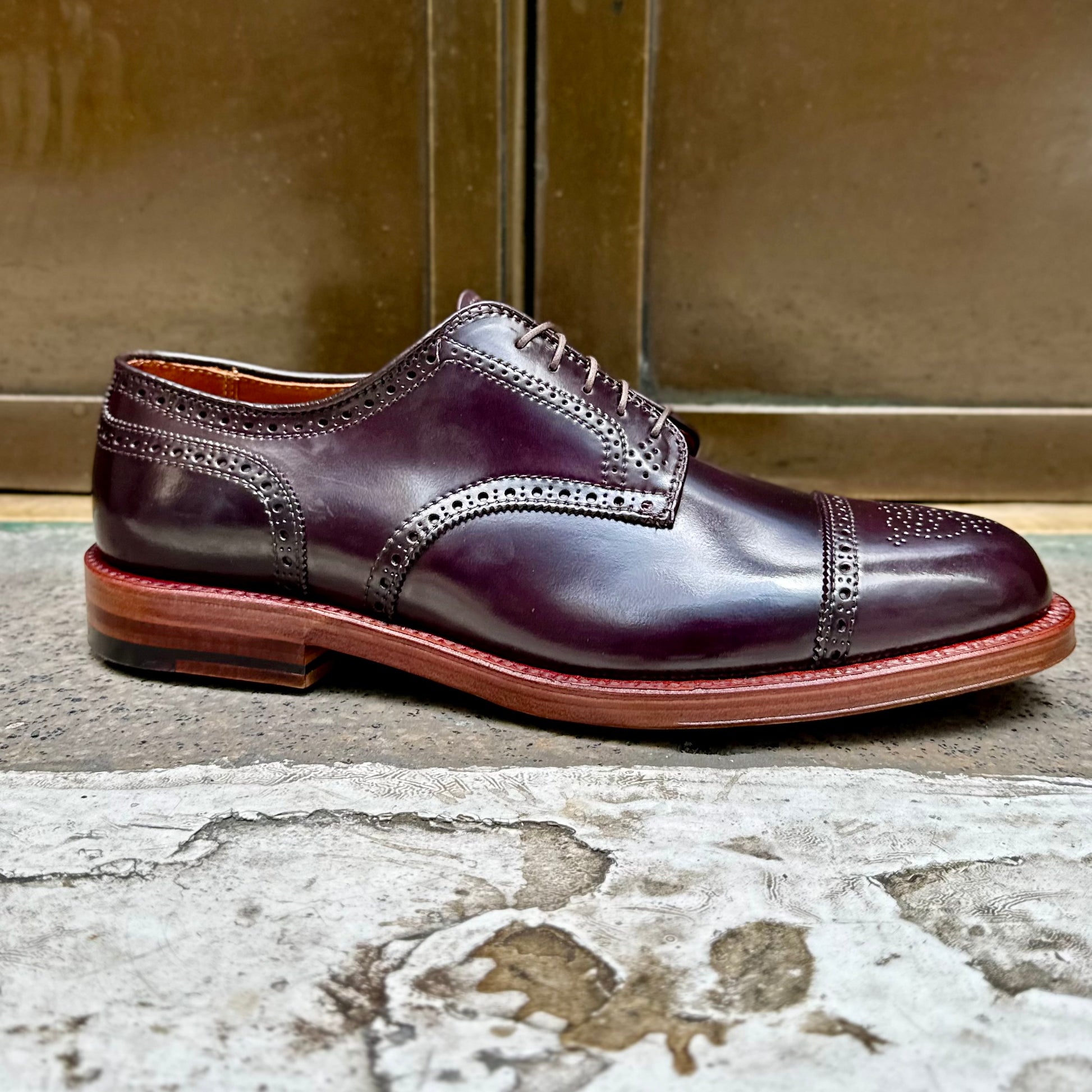 The Alden Madison 21522 - Medallion Cap Toe Blucher Color 8, crafted from Shell Cordovan leather, showcases elegant and durable dark brown polished leather with intricate brogue detailing on the toe cap and sides, against a metallic bronze background on a textured concrete surface.