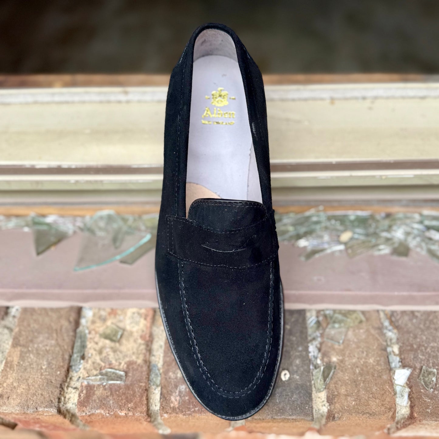 The Alden Madison D2229 “Stuyvesant” Penny Loafer in black suede, with visible stitching and a leather sole, sits on a brick windowsill outdoors among broken glass.