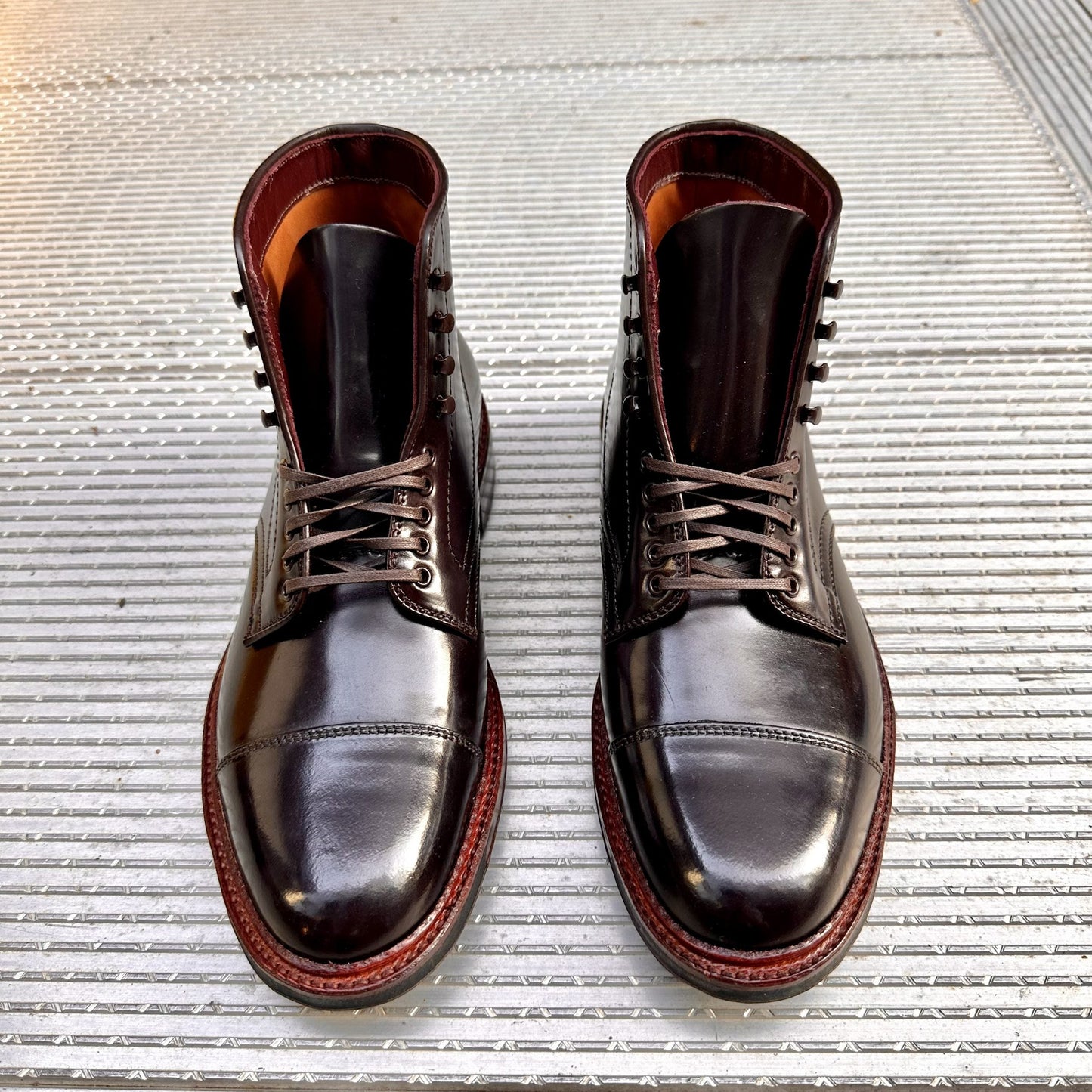 A pair of Alden Madisons D2824HC - Cap Toe Boot w Commando in Color 8 Shell, featuring polished brown leather and laces, is displayed on a textured metal surface. The glossy Shell Cordovan finish highlights their classic and sturdy design as they rest side by side.