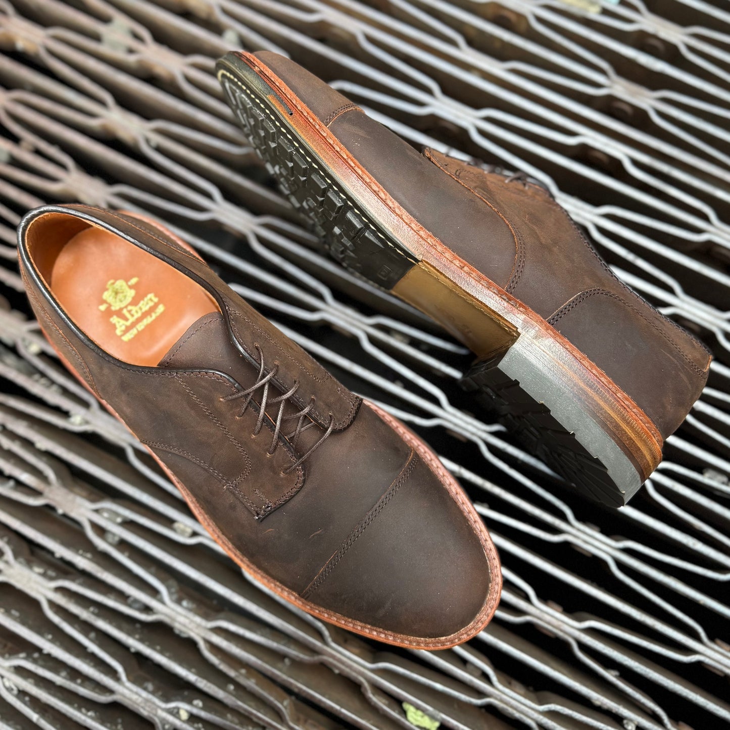 Alden Madisons D4519C - Tobacco Chamois Cap Toe Blucher, brown leather with brown laces, features a durable Commando sole with visible tread. One shoe stands upright while the other shows the brand logo inside on a gridded metal surface.