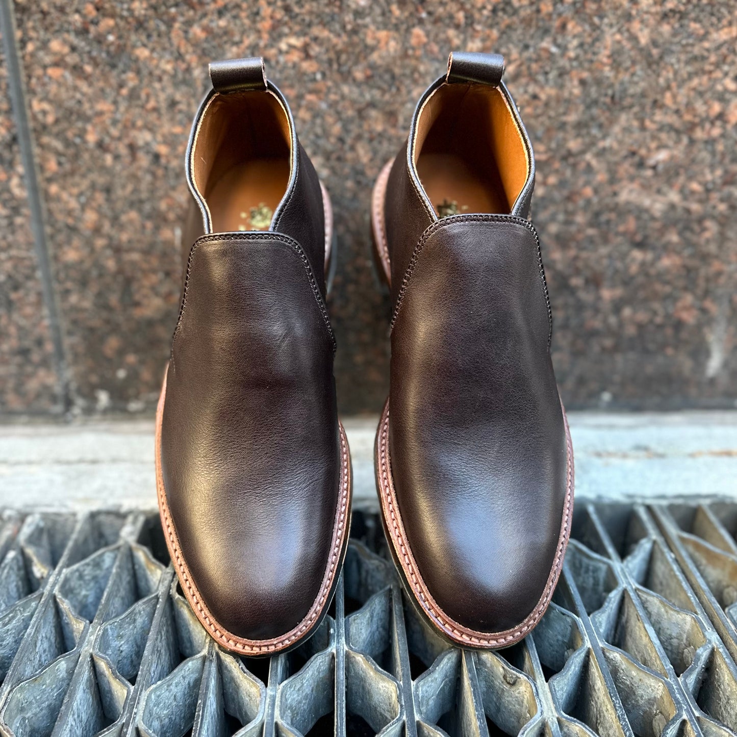 The Alden Madison D2714C - 750 Slip On Chukka in Arabica Lux features dark brown polished leather and light brown Commando Rubber Soles, shown from above on a metal grate with a stone wall background.