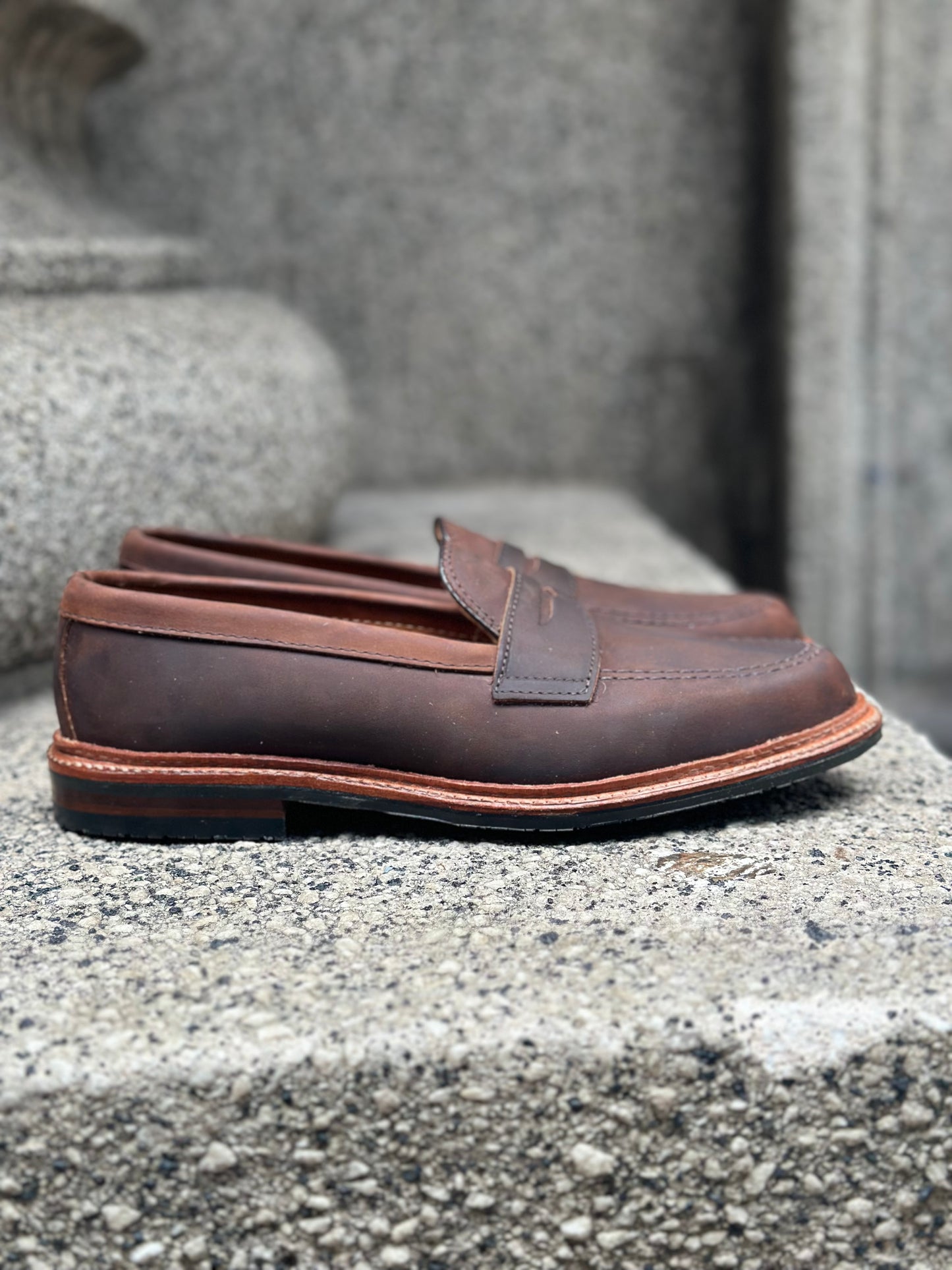 The Alden Madison D2216L - Smooth Tobacco Chamois Penny Loafer, sporting stitched detailing and a durable rubber sole, rests on a textured stone surface with its brown leather finish contrasting against the rough gray background.