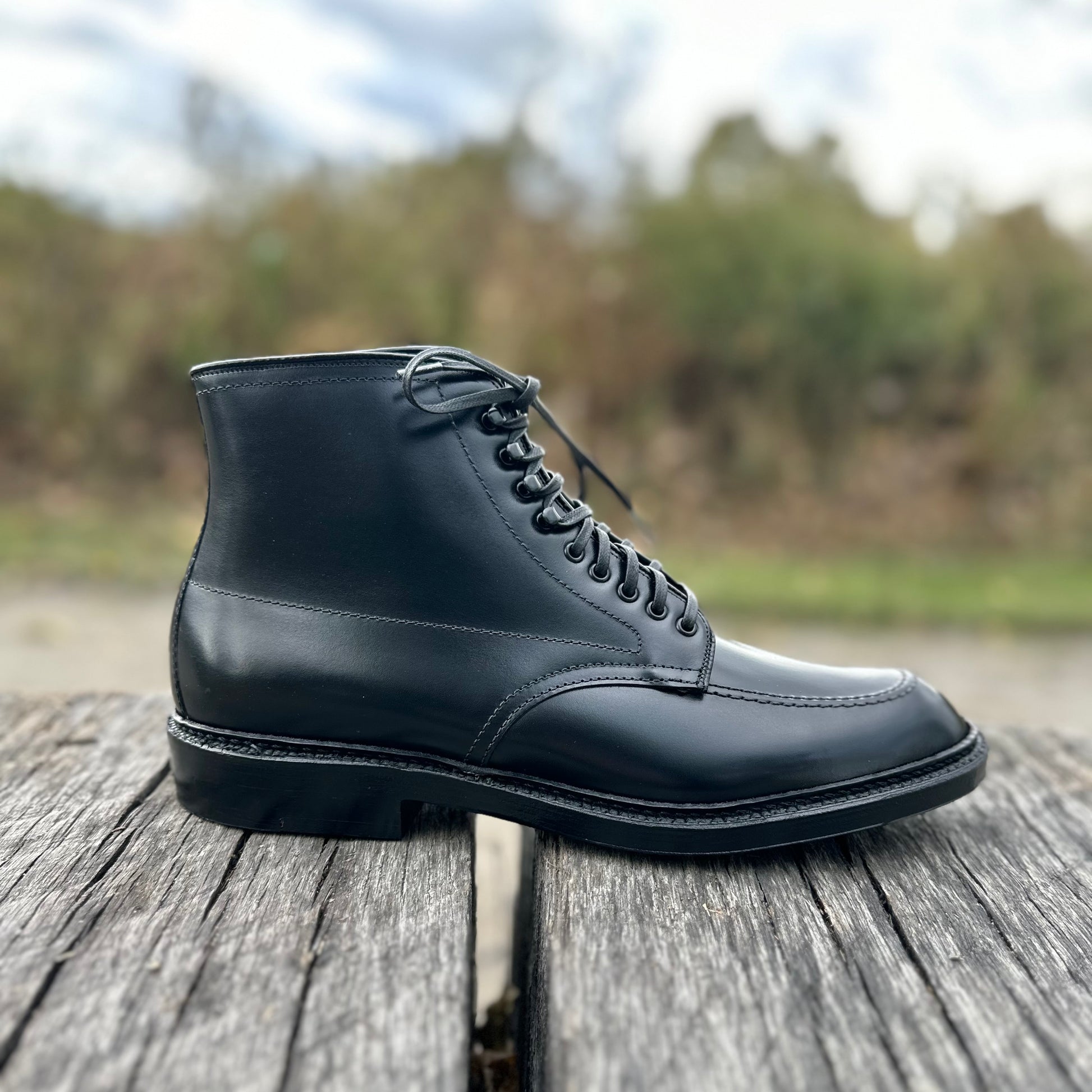 An Alden Madison D2941H - Black Calf Indy Boot with laces sits on a wooden surface outside, against a blurred backdrop of greenery and a cloudy sky.