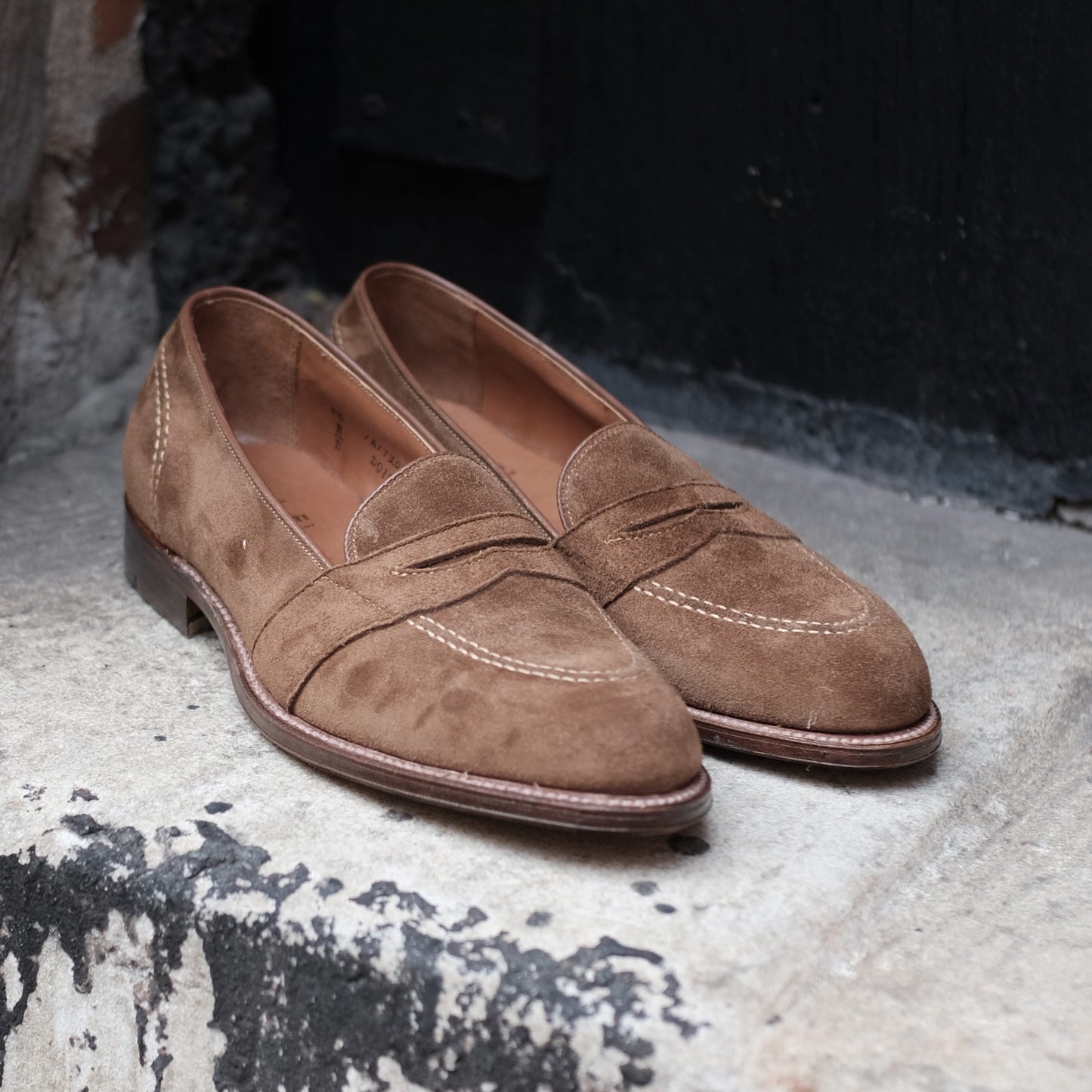 The Alden Madison D0109 Full Strap Loafer in snuff suede features decorative stitching and a leather outsole, shown resting on a weathered concrete step by a dark door and brick wall.