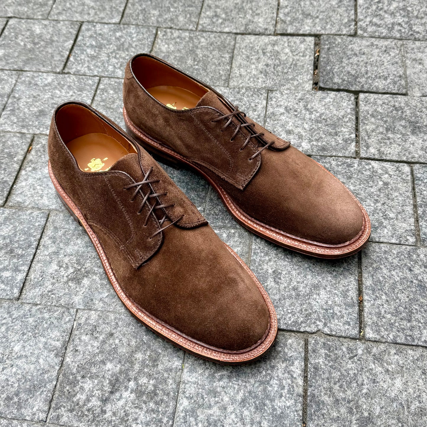 The Alden Madison 29335F - Unlined Dover in Brown Suede, featuring flex leather soles, is showcased on gray stone tile pavement.