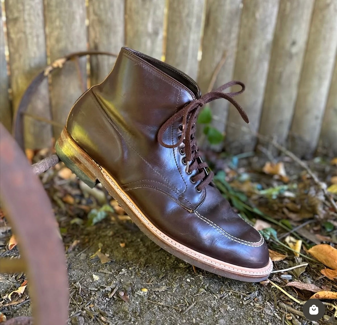 The Alden Madison 403 - Indy Boot in Brown Chromexcel, featuring brown leather, Speed Hooks, and a light sole, stands upright outdoors on dirt with a wooden fence and scattered leaves in the background.