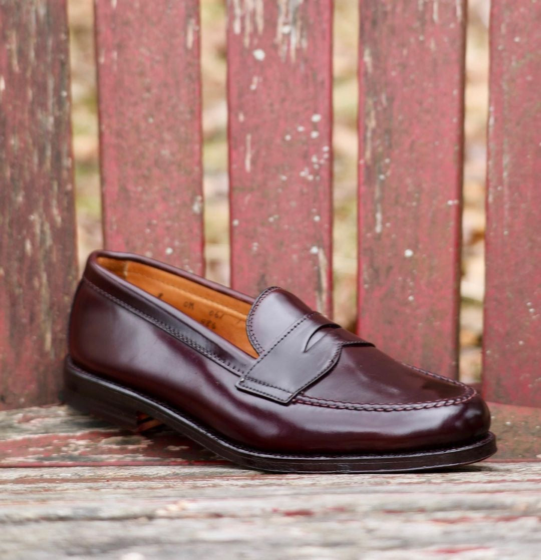 A single 986 - Leisure Hand Sewn loafer in Color 8 Shell Cordovan by Alden Madison sits on a weathered wooden surface with a red backdrop. The polished, visible-stitched shoe is set against a blurred outdoor background of trees.