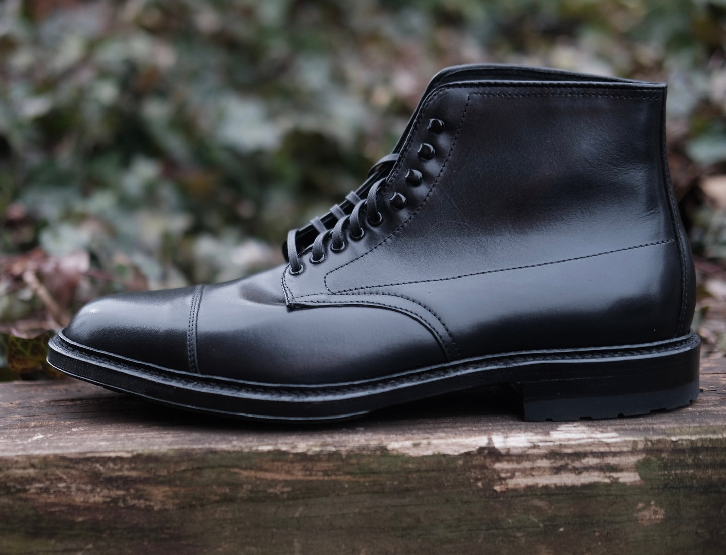 The Alden Madison 40797HC Black Cap Toe Boot with Commando Sole, crafted in black calfskin on the Barrie last, is laced up and displayed outdoors on a weathered wooden surface, set against blurred green foliage.