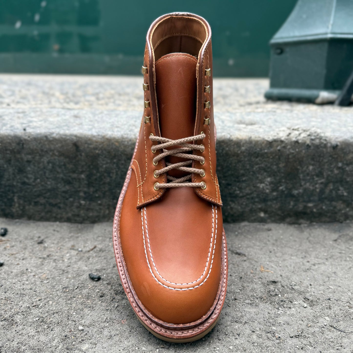 A single Alden Madison Sherwood Indy Boot in Saddle Calf Leather, with visible stitching and beige laces, rests on concrete. A green wall and metal structure backdrop highlight the Cream Wedge Soles rugged elegance.