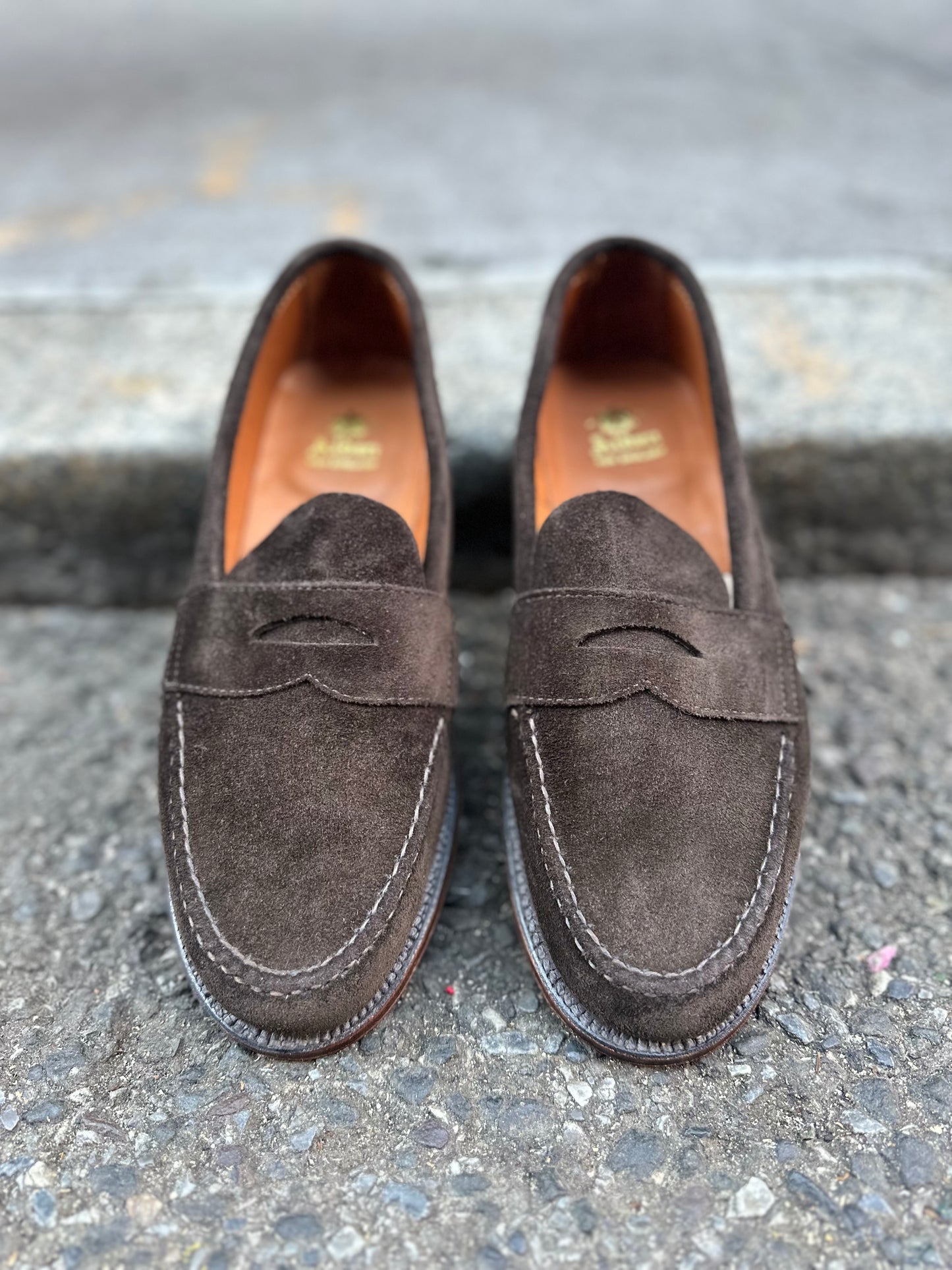 A pair of Alden Madisons unlined leisure hand-sewn loafers in dark chocolate suede rests on a textured stone surface. Designed with decorative cutouts, visible stitching, and a Flex Leather Sole on the Van Last, these shoes offer exceptional comfort and style. Available for preorder.