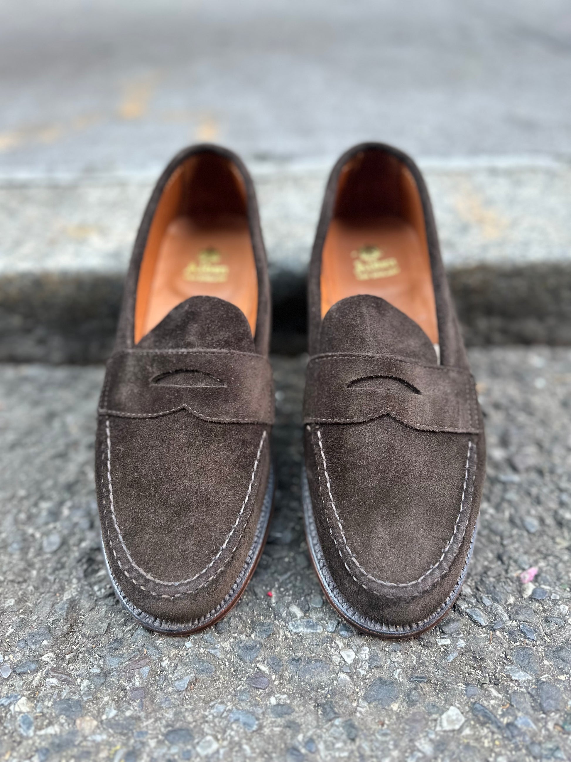 A pair of Alden Madisons unlined leisure hand-sewn loafers in dark chocolate suede rests on a textured stone surface. Designed with decorative cutouts, visible stitching, and a Flex Leather Sole on the Van Last, these shoes offer exceptional comfort and style. Available for preorder.