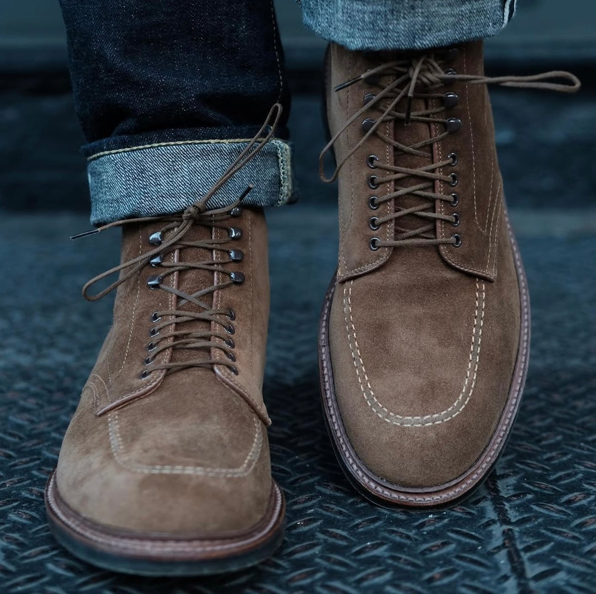 A close-up of a person wearing Alden Madison 4011HC - Indy Boot in Snuff Suede with white stitching and an Antique Commando Sole; cuffed jeans highlight the boots clearly on a textured dark surface.