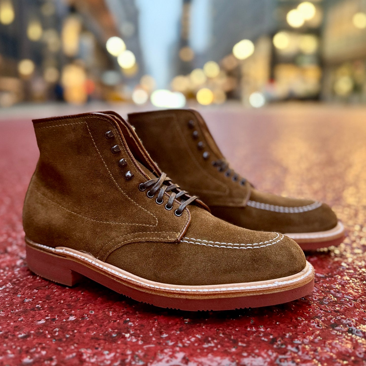The Alden Madison 40531H - Redwood Indy Boot in snuff suede, with detailed stitching, sits on a red textured surface. The background is an out-of-focus urban street scene, highlighted by bright lights, suggesting evening or dusk.