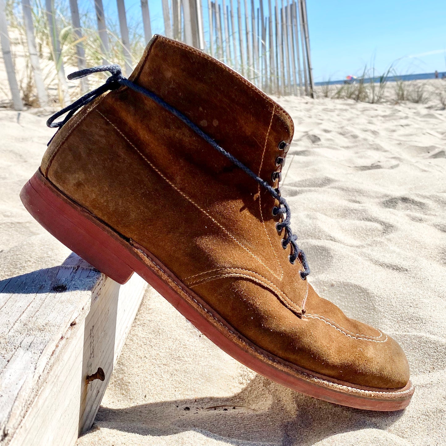 A 40531H - Redwood Indy Boot in Snuff Suede from Alden Madison with black laces rests on a wooden rail, its red rubber sole glows in the sun against a sandy backdrop with a beach fence under a clear blue sky.