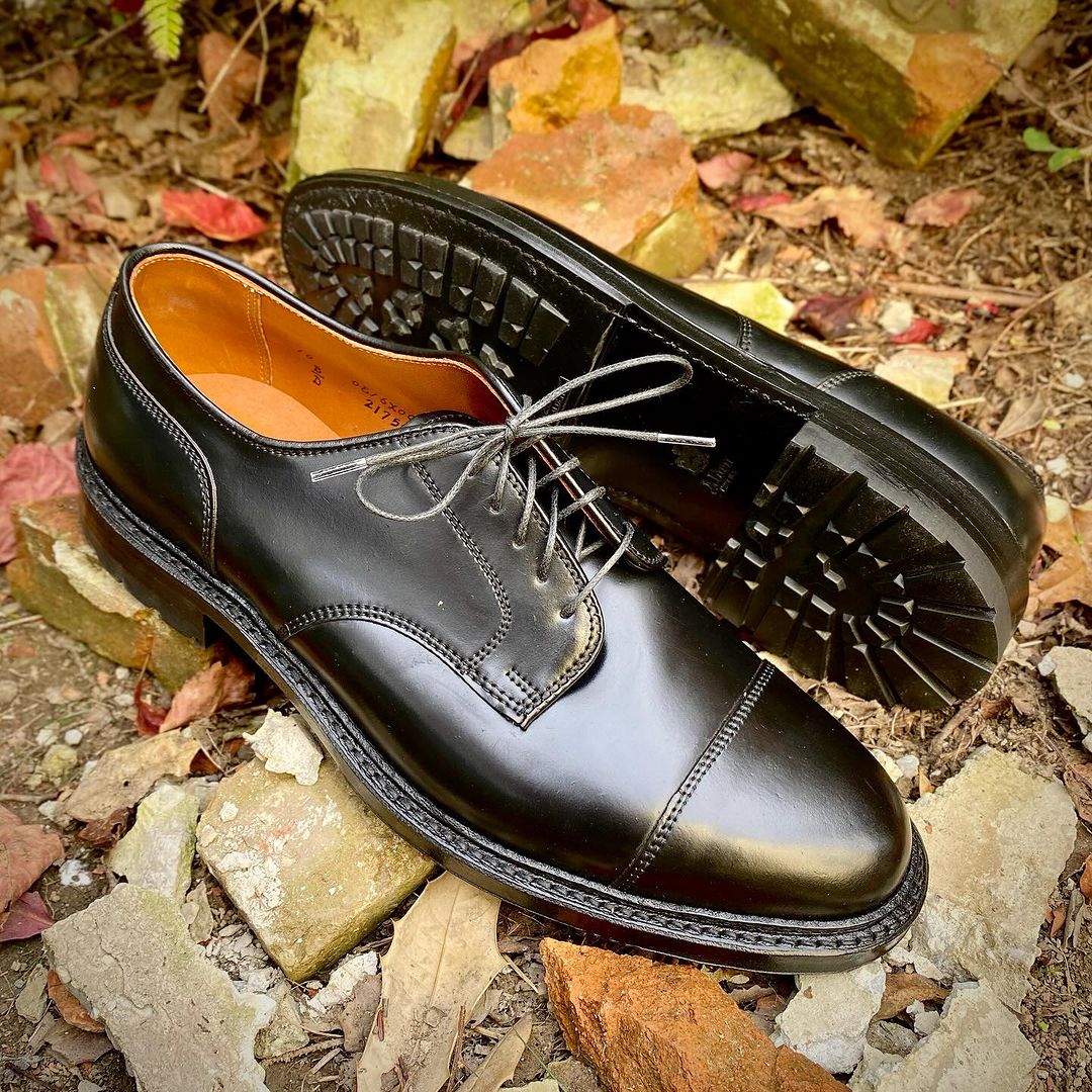 A pair of Alden Madison 2175C - DeRosa Captoe Blucher shoes in polished black shell leather with brown interiors and sturdy Commando soles sits on a stone- and leaf-covered ground.