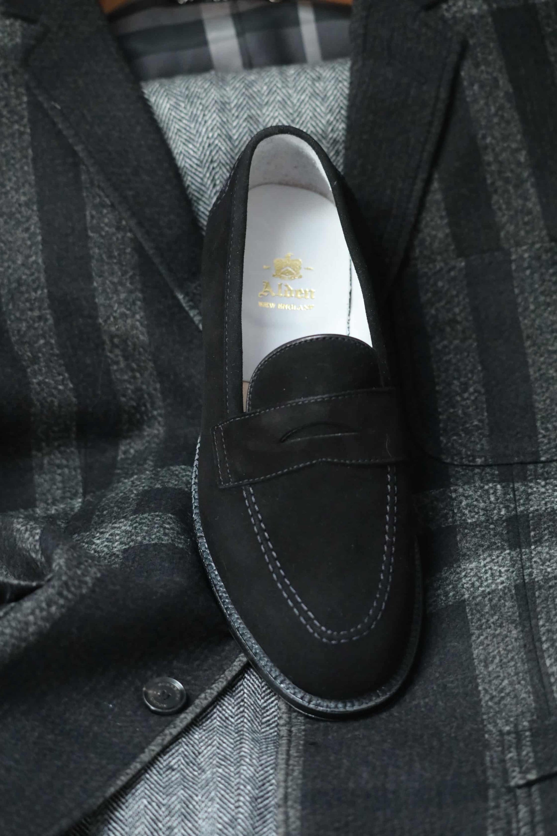 The D2229 “Stuyvesant” Madison Penny in black suede by Alden Madison, with white stitching and a sleek leather sole, is displayed atop a dark gray plaid blazer with large checks and buttoned front.