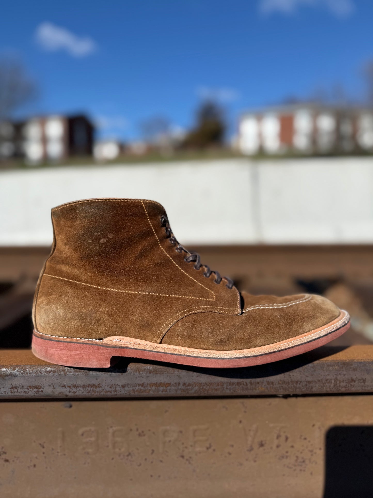 The Alden Madison 40531H - Redwood Indy Boot in Snuff Suede, featuring a red rubber sole, rests on a metal rail with blurred residential buildings and a clear sky in the background.