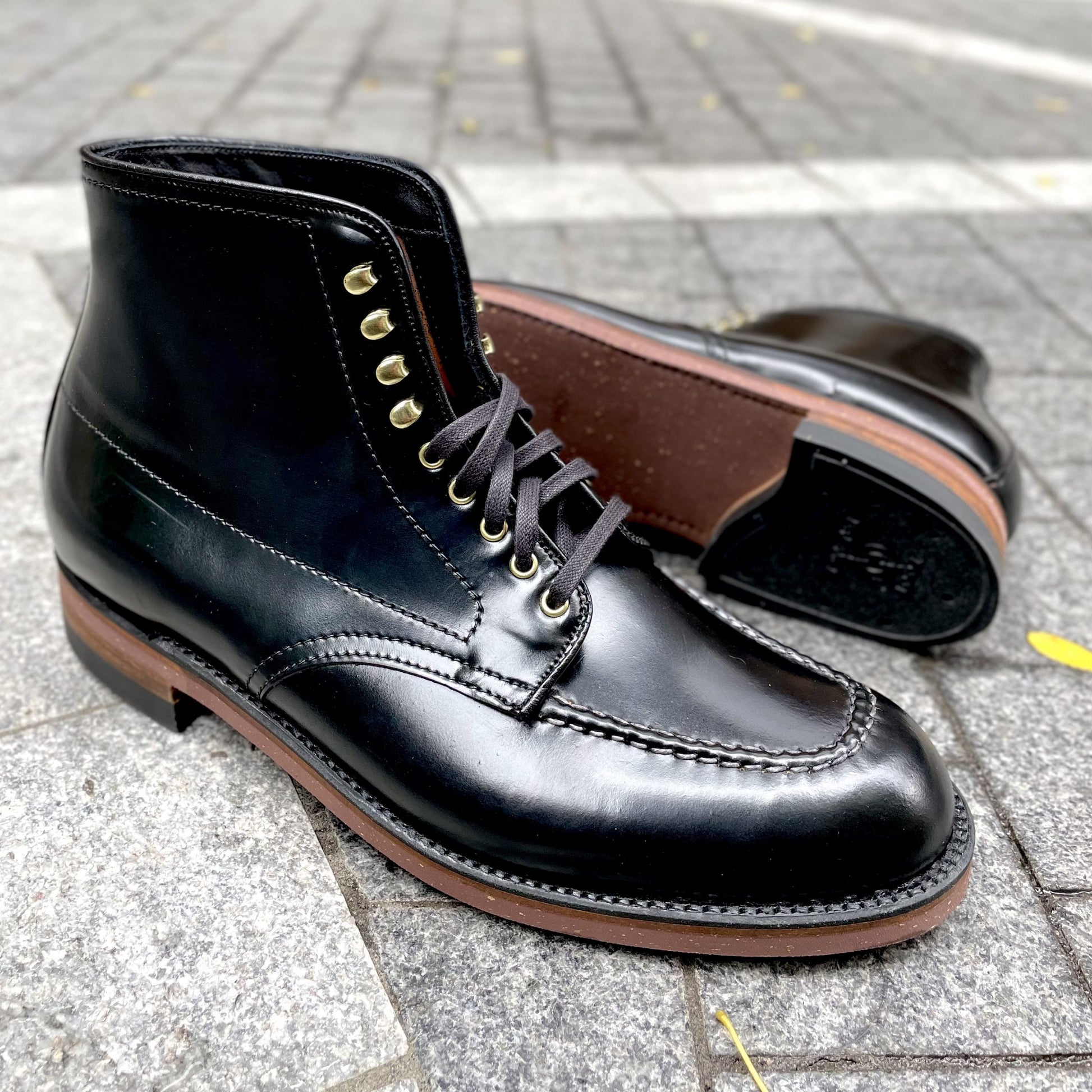 Alden Madisons D1908H - Copake Indy Boot in Black Shell features brown soles and gold eyelets, with one shiny black boot standing upright and the other lying on its side on gray stone pavement.