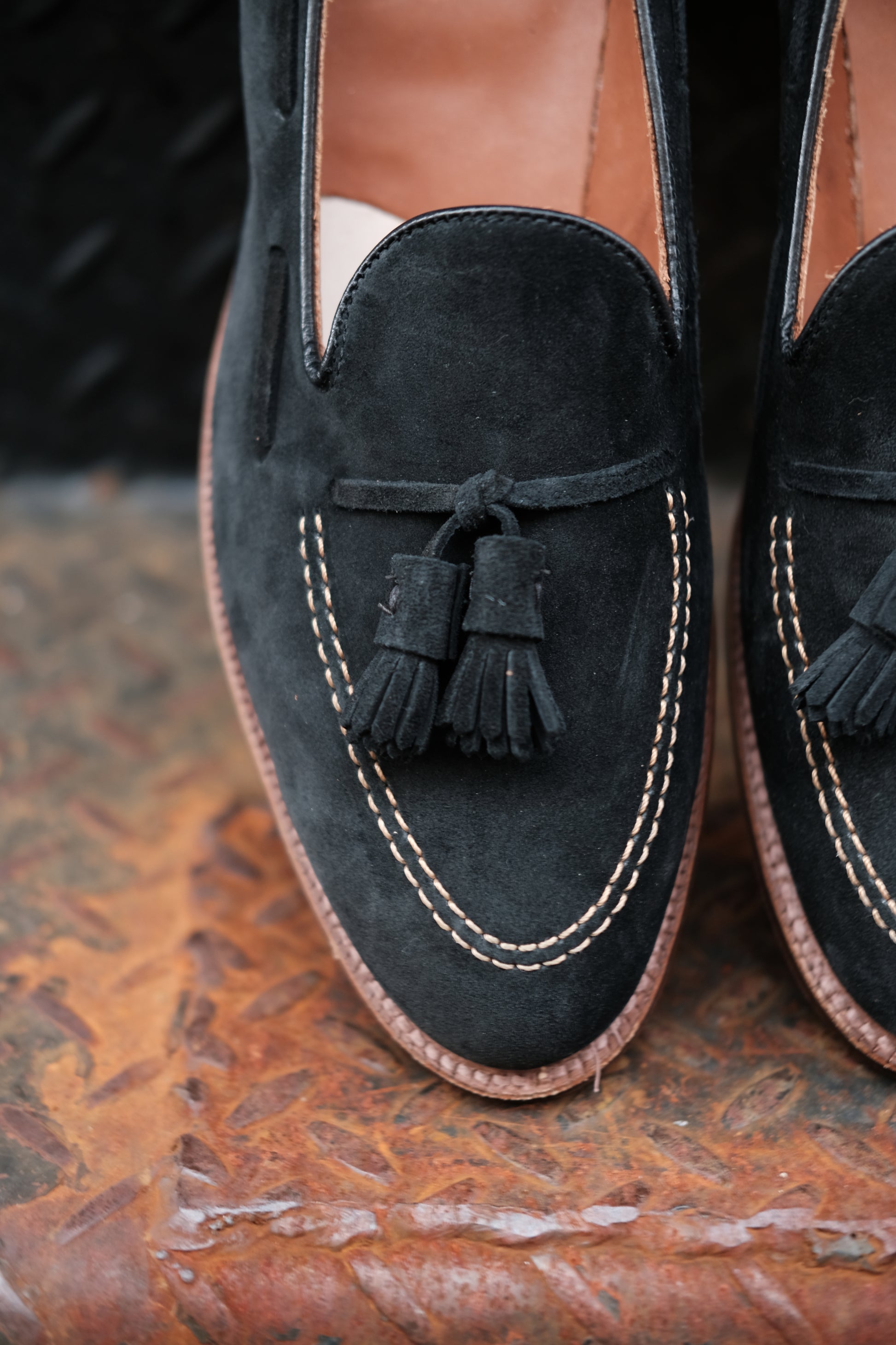 Close-up of Alden Madison D0106 - Black Suede Tassel Loafer, crafted on the Aberdeen Last with tan stitching and decorative tassels, displayed on a textured rust-hued metal surface with visible sole edges.