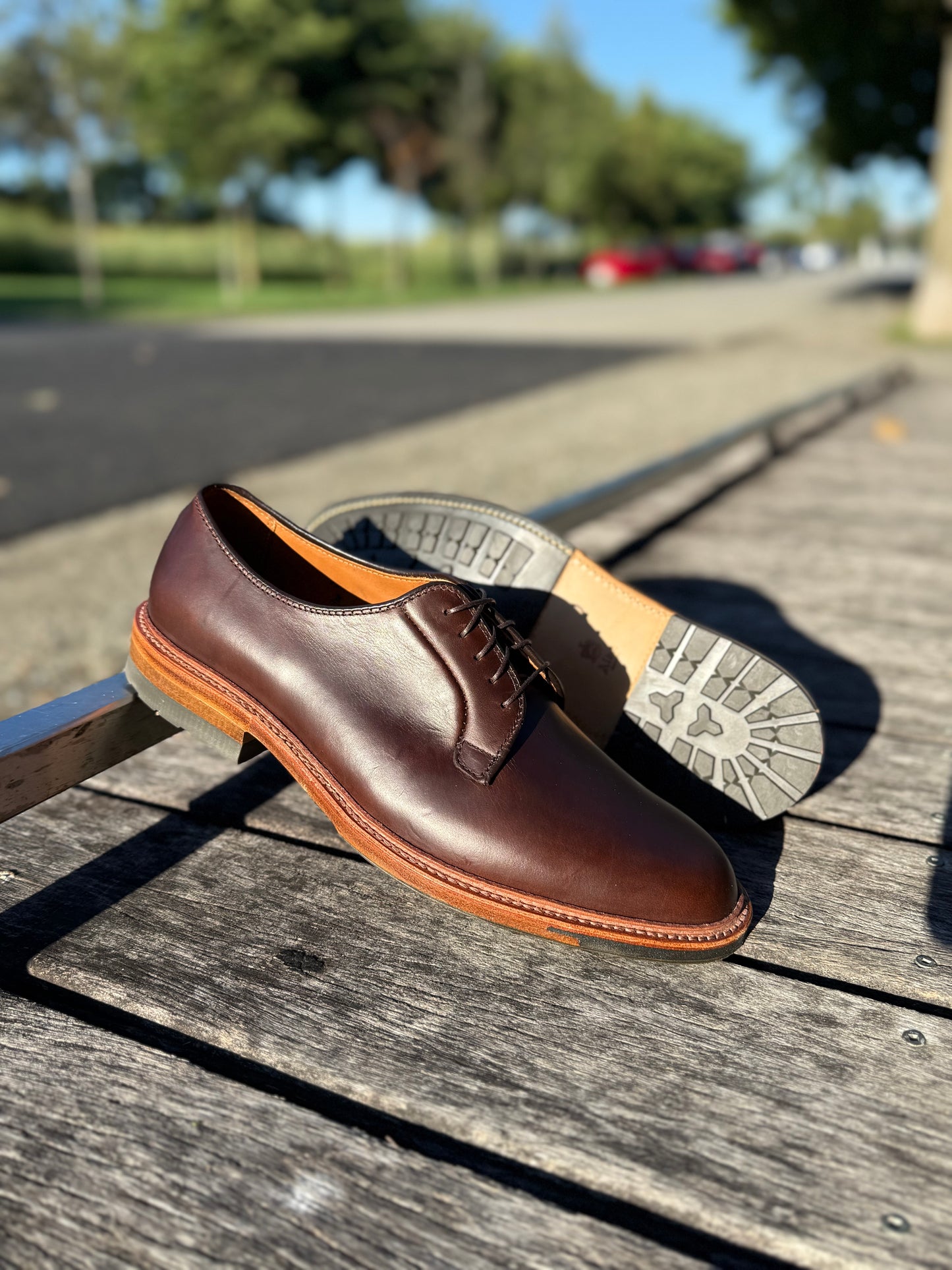 A pair of ALDEN SHOES 9506C - Plain Toe Blucher w Commando Brown CXL is displayed on a wooden bench outdoors, with trees, grass, and a road softly blurred in the sunny background.