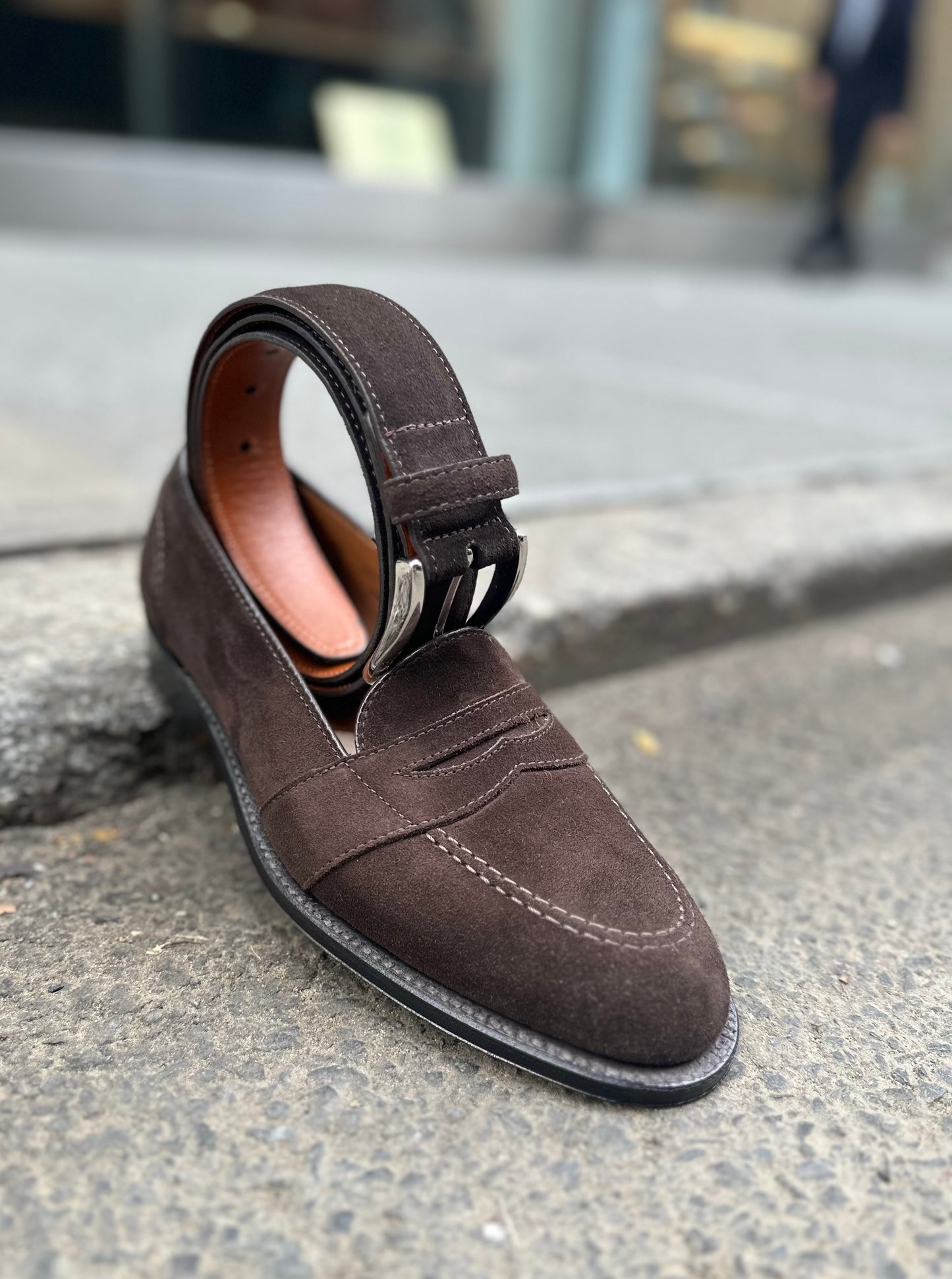 The ALDEN SHOES G9201 - Dark Brown Suede Full Strap Loafer with a silver buckle sits on a concrete curb, set against a blurred urban sidewalk and building backdrop.