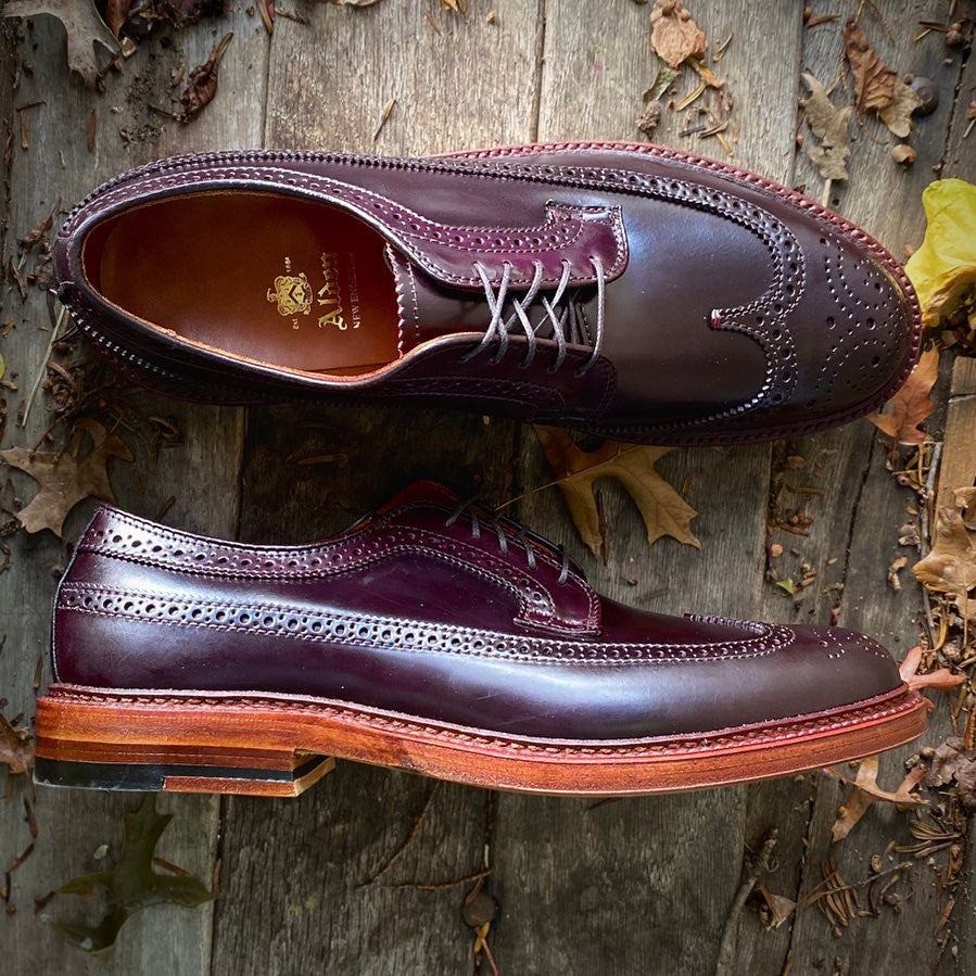 The Alden Madison D5511 Antique Long Wing Blucher in Color 8 Shell Cordovan, featuring brogue detailing, brown laces, and double leather soles, is displayed on a rustic wooden surface with autumn leaves scattered around.