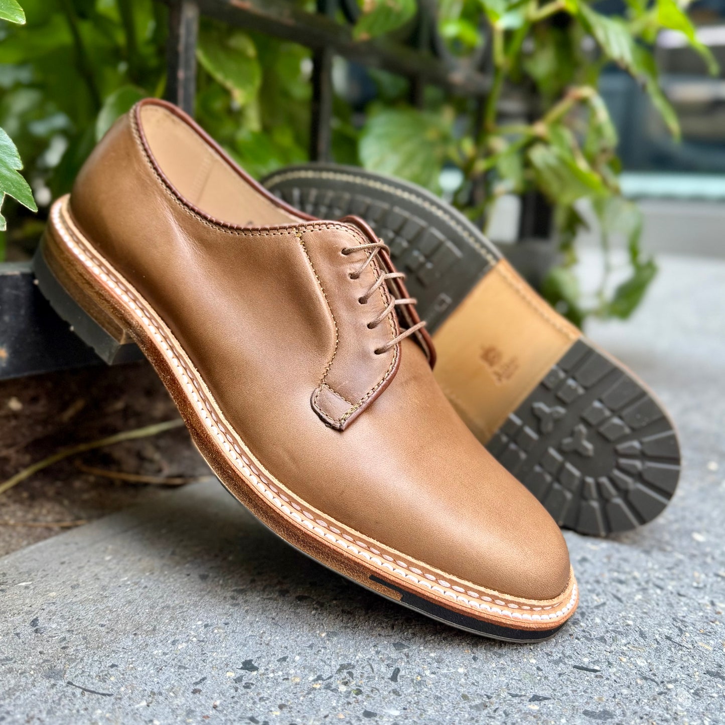 The ALDEN SHOES 9501C Natural CXL Plain Toe Blucher in tan Chromexcel leather, with brown laces and a black rubber sole, is shown outdoors on stone with greenery in the background, one shoe displayed on its side.