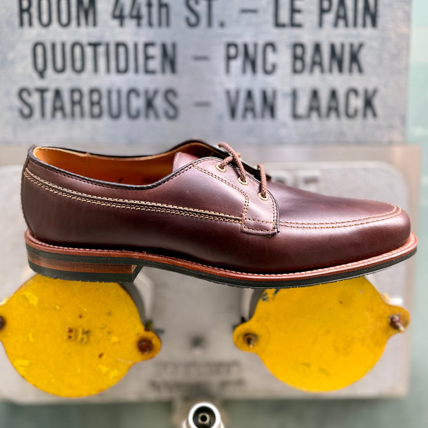 The Alden Madison 734L - Ivy Blucher in Brown Chromexcel, a leather lace-up dress shoe, rests atop two yellow circular metal fixtures; behind it, a blurred sign lists various businesses.