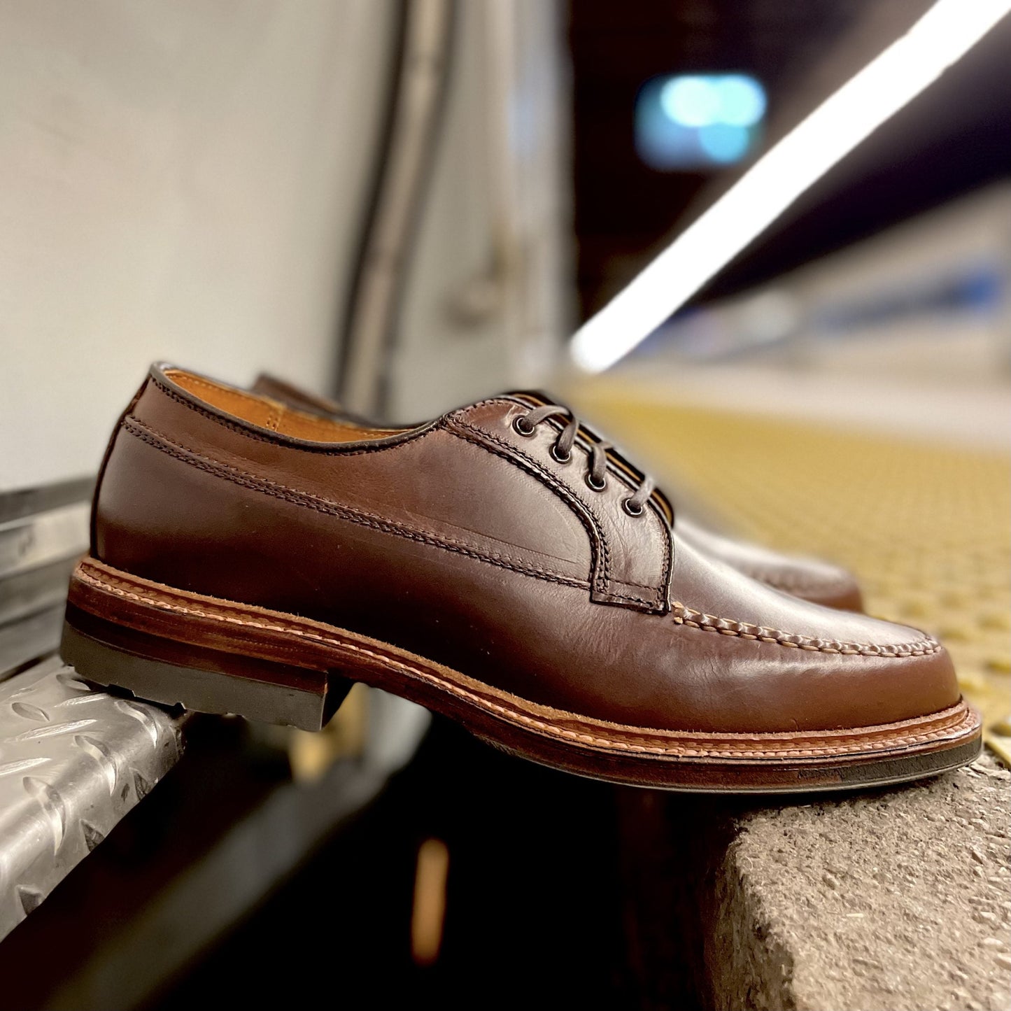 The Alden Madison D1611C - Ranger Mocc in Brown CXL, featuring laces, an antique edge, and detailed stitching, rests on a metallic surface near a train platform edge amid a blurred background.
