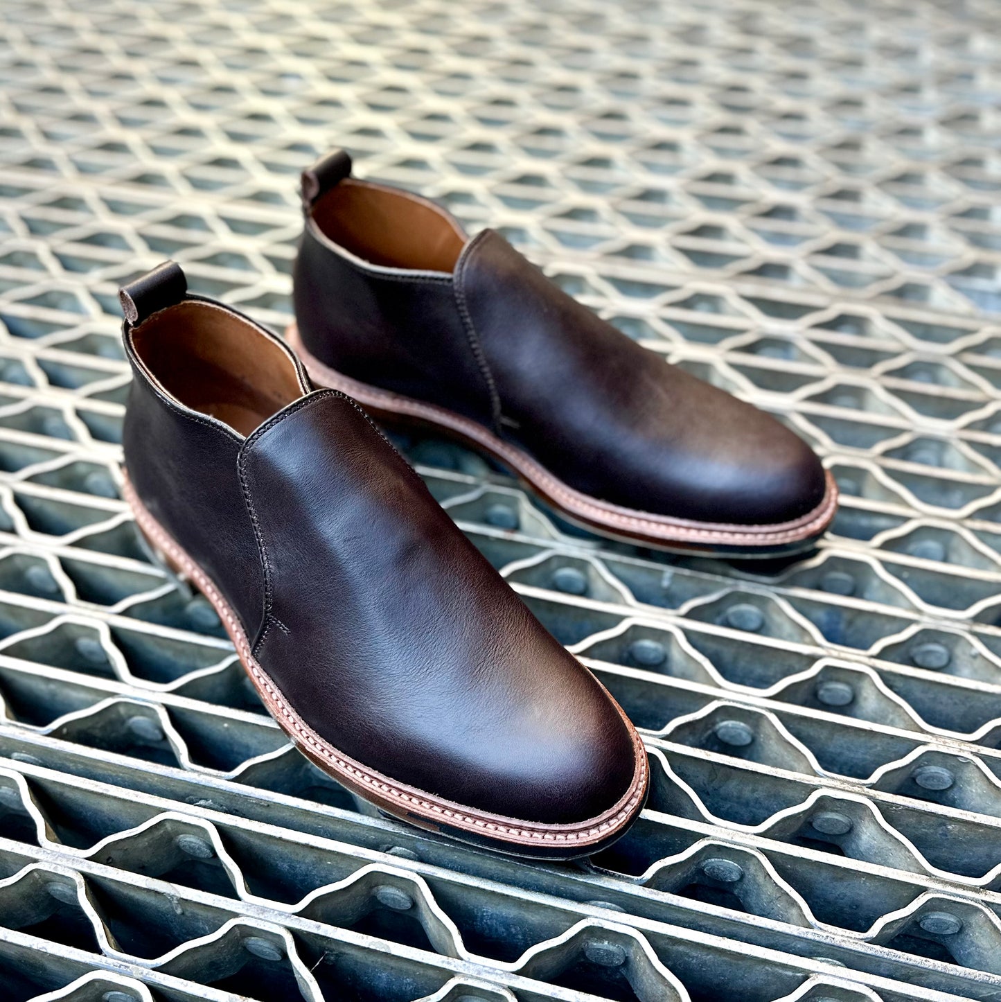 The Alden Madison D2714C - 750 Slip On Chukka in Arabica Lux, featuring dark brown leather and tan Commando rubber soles, is displayed side by side on a geometric-patterned metal grate, facing slightly to the right.
