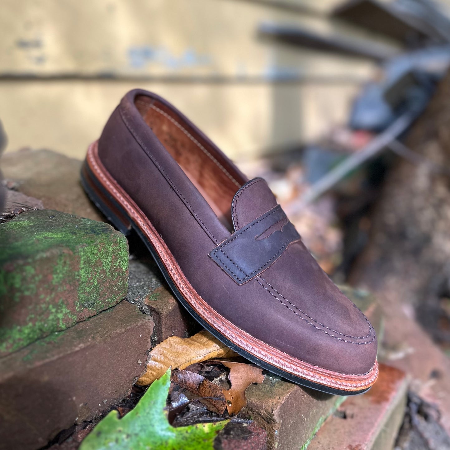 An Alden Madison D2216L - Smooth Tobacco Chamois Penny Loafer with a stitched rubber sole sits on aged, mossy bricks. Fallen leaves and weathered outdoor elements give an antique feel to the scene.