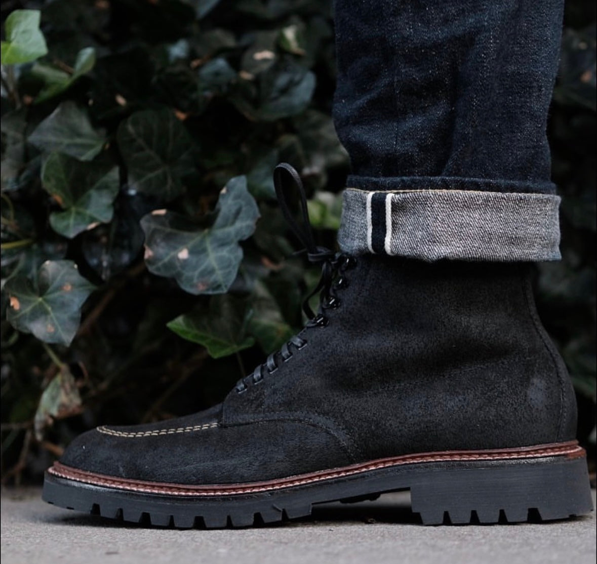 A person wearing dark cuffed jeans and Alden Madison D9916H Indy Boot in Earth Reverse Chamois with a Vibram Heavy Lug Outsole stands on concrete, green leafy plants in the background.