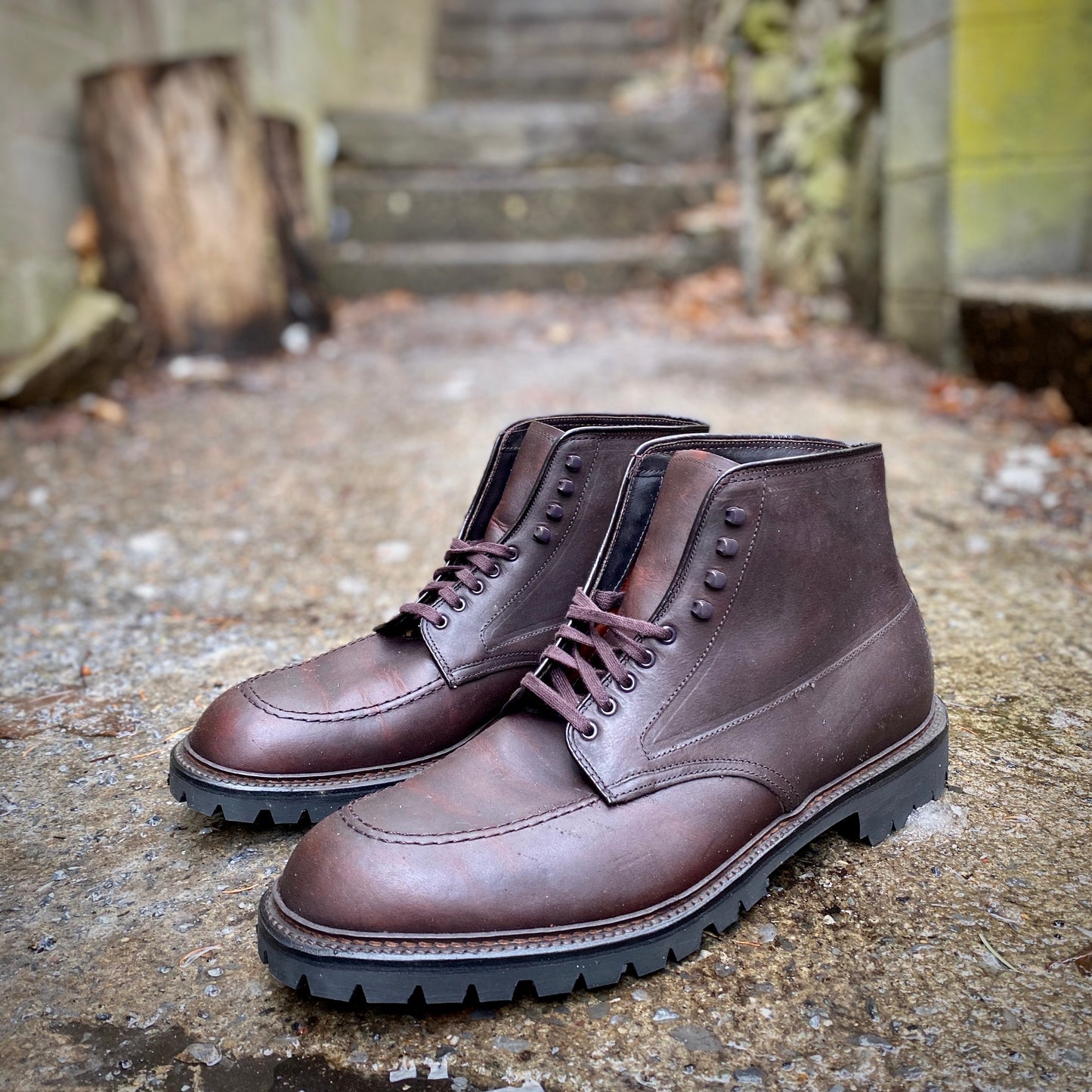 The Alden Madison 404 - Indy Boot in Kudu (Oiled CXL), with Vibram lug outsoles, sits outdoors on a concrete surface. Mossy stone steps and a weathered wooden post create a rustic backdrop.