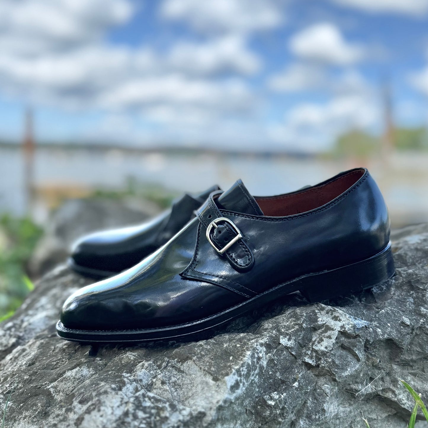 Alden Madison’s 1655 - Monk Strap Black in Shell Cordovan, featuring silver buckles and a single oak leather sole on the Aberdeen Last, sits atop a gray rock with blue sky, clouds, water, and greenery in the blurred background.