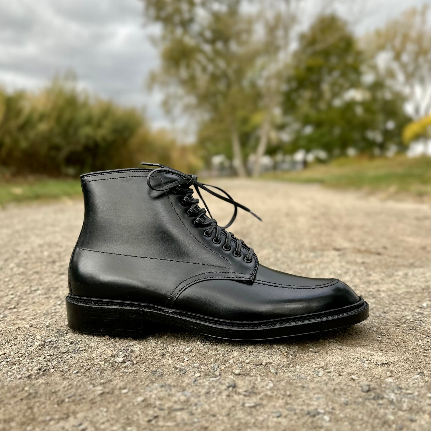 A single Alden Madison D2941H - Black Calf Indy Boot with a double leather sole is set on a gravel path. In the background, blurred green trees and a cloudy sky enhance the serene outdoor atmosphere.