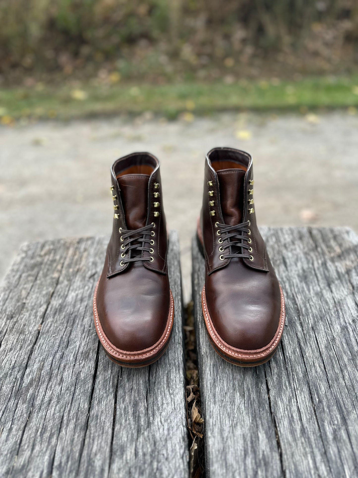 A pair of Alden Madison D1848H Plain Toe Boots in Brown Chromexcel, featuring gold eyelets and a crepe rubber sole, rest side by side on a weathered wooden surface outdoors with grass and pavement blurred in the background.