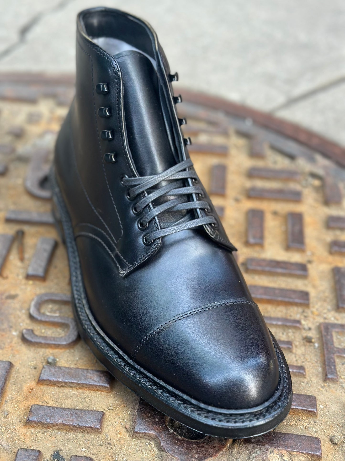 The Alden Madison 40797HC Black Cap Toe Boot with Commando Sole is shown outdoors on a metal manhole cover.