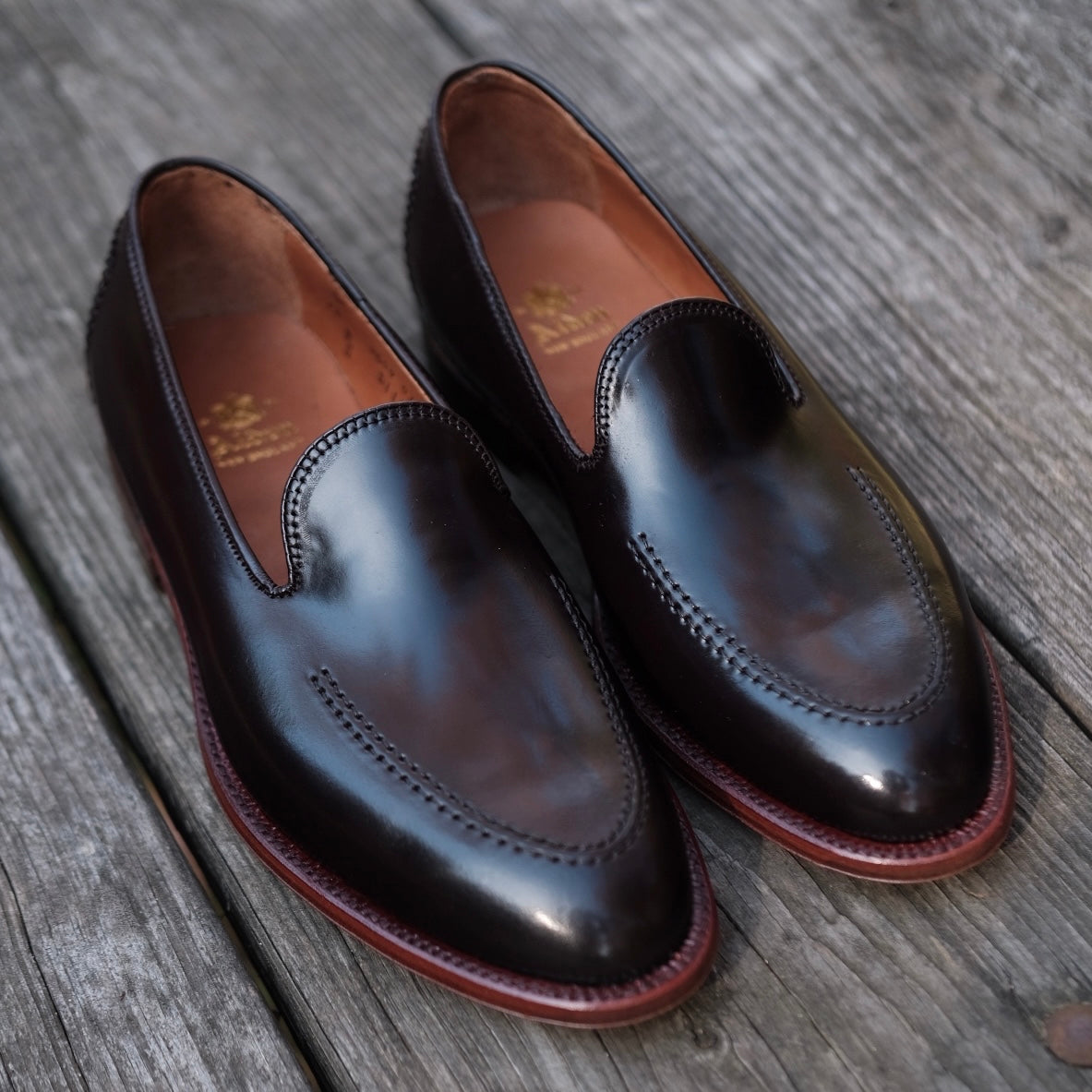 The Alden Madison Vanderbilt Slip On (D1118) in Color 8 Shell, designed on the Aberdeen Last with sleek stitching and a polished finish, sits on a weathered wooden surface.