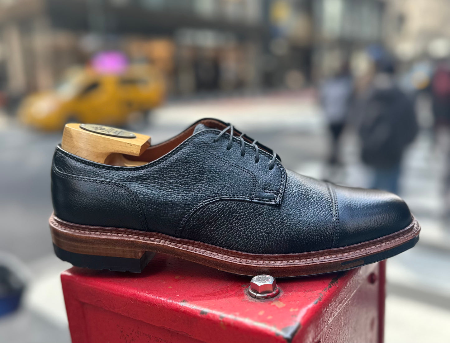Displayed on a red surface in an urban setting, the Alden Madison 22826C - Black Regina Grain Derosa Blucher with a wooden shoe tree stands out, contrasting against blurred pedestrians and a yellow taxi.