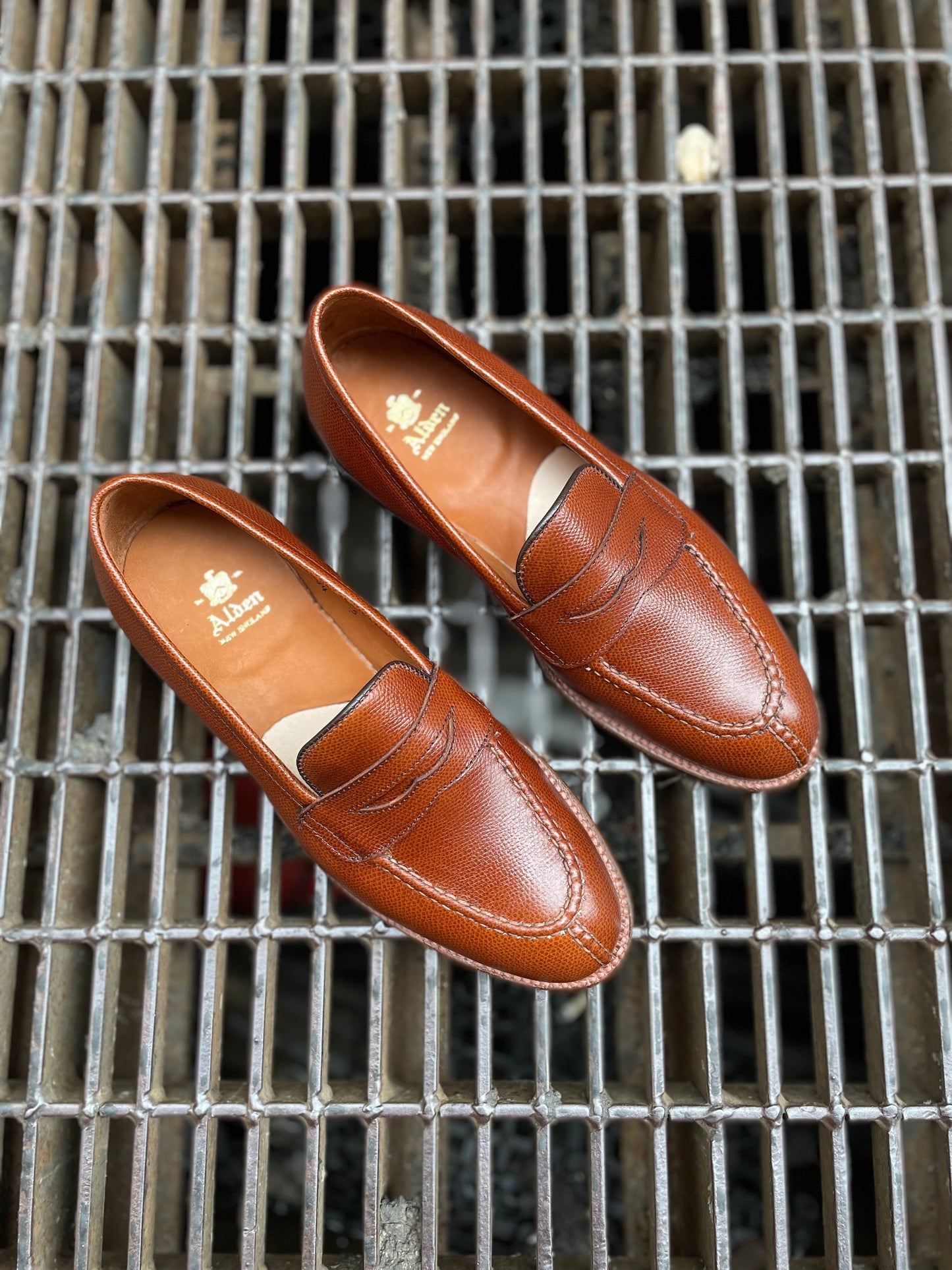 A pair of Alden Madison D1204 - Alpine Grain NST Loafers in brown leather with a polished finish, visible stitching, and gold Alden branding on the insoles rests on a metal grate.