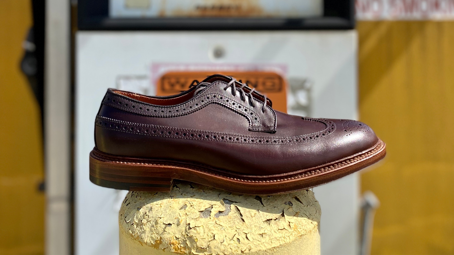 The Alden Madison 976 - Long Wing Blucher in Brown Calfskin, featuring detailed brogue patterns and a double waterlock sole, rests outdoors on a rugged concrete surface. A vintage gas pump and yellow wall can be seen blurred in the background.
