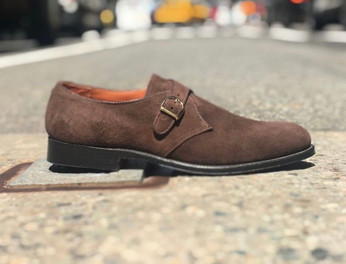 The Alden Madison 953 - Monk Strap in Brown Suede, featuring a leather sole and side buckle, is displayed on a city street with blurred cars and buildings in the background.