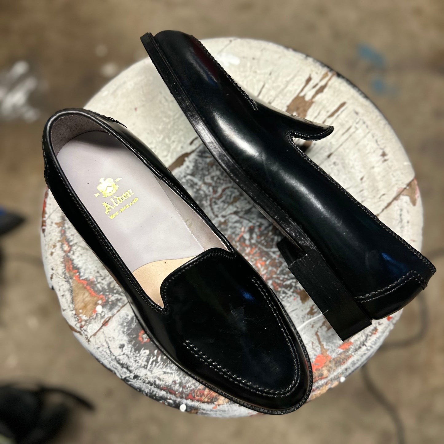 A pair of Alden Madisons D2117 - Vanderbilt Slip On in Black Shell Cordovan loafers sits on a round, paint-spattered stool. One shoe is resting on its side revealing the single oak leather sole, while the other stands upright, displaying the interior branding.